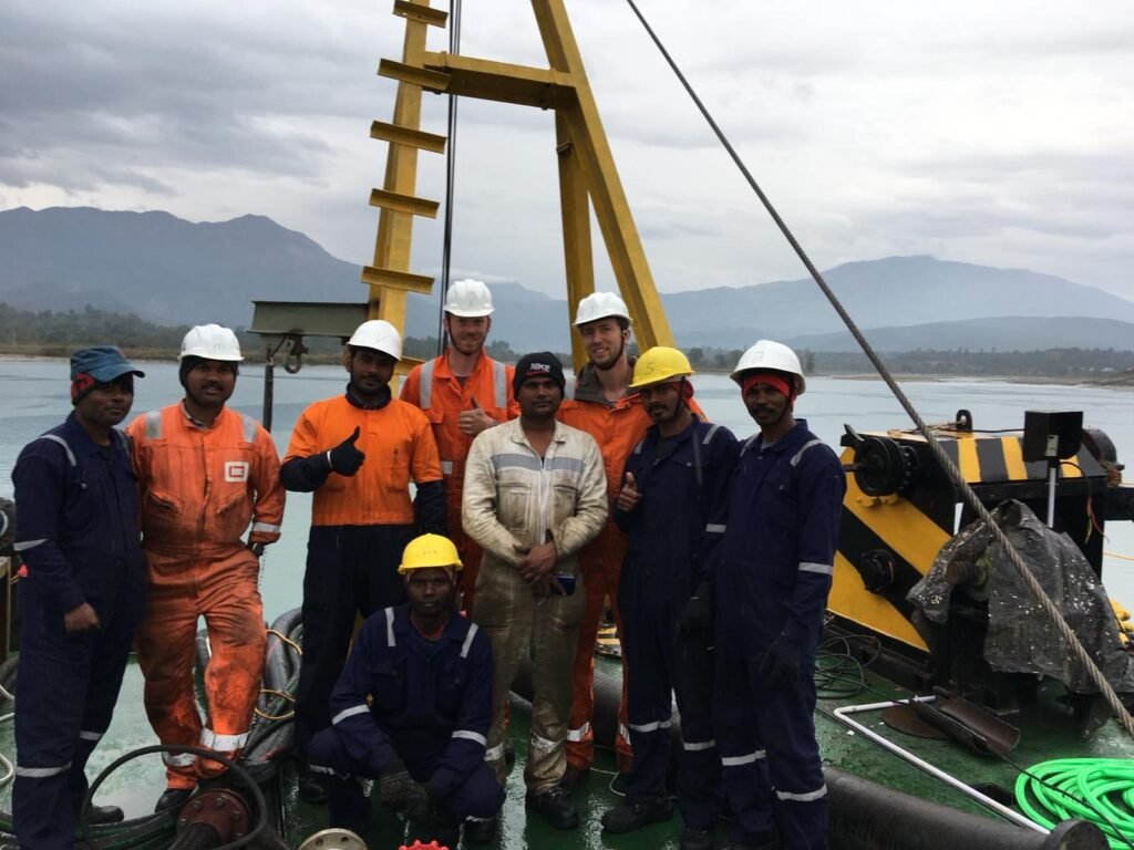 Group of engineers and workers in safety gear posing on equipment at Dakpathar Barrage, Uttarakhand with mountains in the background