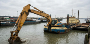 Backhoe dredger on a pontoon performing precision dredging work in a harbor, removing compact seabed material as part of marine engineering and dredging operations.